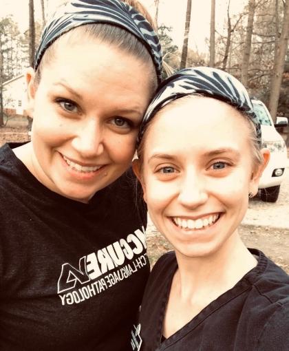 In Her Sister’s Steps - Two women wearing patterned headbands smile in an outdoor selfie. They stand close together in a wooded area with trees and a parked vehicle behind them. One woman wears a black T-shirt with white text, while the other wears a black top. Both face the camera and appear cheerful.