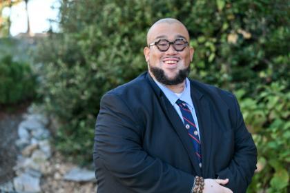 A person stands outdoors smiling, wearing a dark suit, white shirt, and striped tie. Trees and greenery fill the background, with sunlight creating a soft natural light.