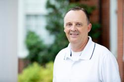 - Man with short dark hair smiles while standing outdoors on campus. He wears a white polo shirt with dark trim along the collar. Green foliage and a brick building appear softly blurred in the background. The portrait is taken in natural light with the subject facing the camera.