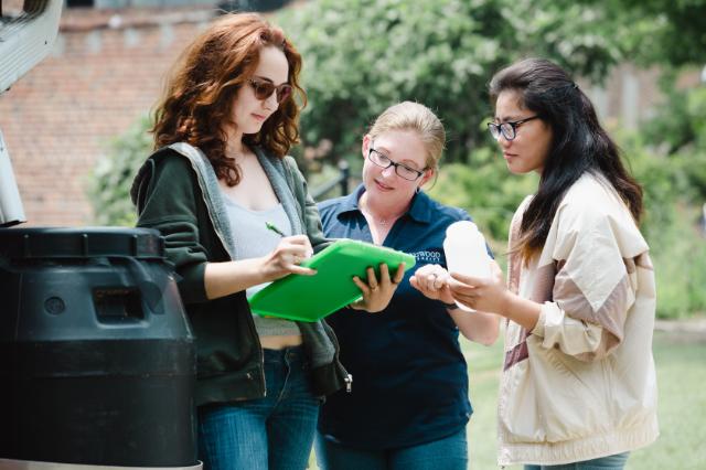 - Three students stand outdoors examining a sample container during a field activity. One student writes notes on a green clipboard while another holds the sample bottle. A black collection bin sits nearby as part of the environmental work. Trees and campus buildings appear in the background on a sunny day.