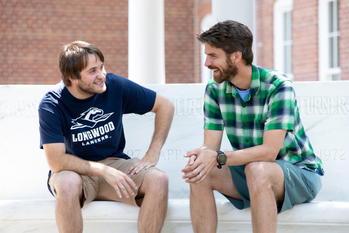 Two people talk on the steps of Chichester - Two men sit on a stone bench outside on campus talking and smiling. One wears a navy Longwood Lancers T-shirt and shorts, while the other wears a green plaid shirt and shorts. They lean toward each other in conversation in front of a brick campus building with white columns. The scene shows a relaxed conversation between members of the Longwood communit