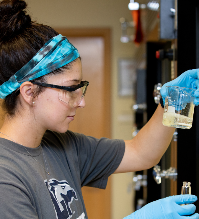 Student looks at beaker in Science Lab - A student wearing safety glasses and gloves examines a beaker containing liquid in a chemistry lab. She holds the beaker up at eye level while working at a fume hood. Her other hand steadies a small glass vial beneath the workstation. The scene shows careful observation during a hands-on chemistry experiment.