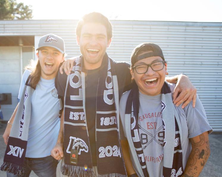 Students with Longwood scarves - Three students stand close together smiling with arms around each other. They wear Longwood scarves and casual clothing outdoors in bright sunlight. One student wears a Longwood cap while another wears glasses and a backwards hat. Friends celebrate together during a campus event.