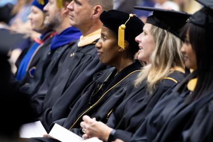 Faculty at convocation - Faculty members sit in a row during a campus convocation ceremony. Several wear black academic gowns and doctoral hoods. One faculty member wears a black tam with a gold tassel and looks toward the stage. Programs are held in their laps as the audience blurs in the background.