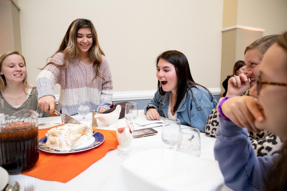 Students cuts the Baked Alaska while her fellow guests watch - Student slices a meringue-topped dessert on a platter at a dining table. Friends lean in smiling and laughing as the dessert is served. A pitcher of iced tea, water glasses, and orange napkins sit on the table. The group is seated together in a dining hall.