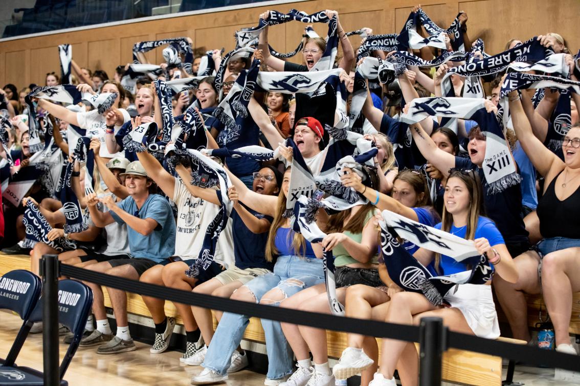 Students with Longwood scarves - Crowd of Longwood students cheer from the bleachers inside a gym arena. Many hold blue-and-white “We Are Longwood” scarves with the Lancer logo. Students shout and raise scarves overhead during the spirit event. Signs reading “The G.A.M.E. XIII” appear among the crowd.