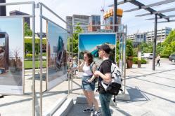 Students looking at art on the street - Two students view large photo prints displayed in an outdoor exhibit. One student points toward an image while both stand beside the metal display frames. Bright midday sunlight casts long shadows across the plaza. Downtown buildings and a construction crane rise in the background.