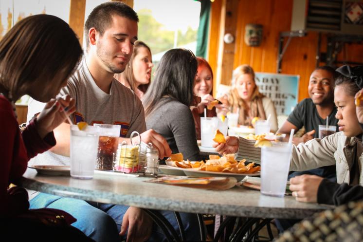 Students at Macados in Farmville - Group of students sits around a table eating chips and salsa at a restaurant. Water cups and soft drinks sit on the table as people talk and smile. Warm wood-paneled interior and daylight from windows frame the scene. Hands reach for chips while conversation continues across the table.