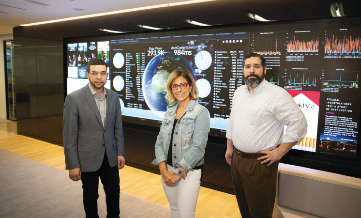 Travis Lyles '15 (left) with Anthony J. Rivera '03 and Anna Knapp '97 who also work at The Washington Post [Photo from 2019] - Three professionals stand in front of a large digital analytics dashboard display. The screen shows charts, graphs, a world map, and website performance metrics. One person wears a gray suit, another a denim jacket, and another a white shirt. They pose in a modern office s