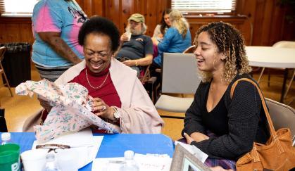 Sylvia Maxine Adamson and Sjory Traverso '24 - An older woman smiles as she opens a wrapped gift at a table. A younger woman sits beside her, laughing and watching. They are seated in a wood-paneled room with other people in the background. Papers, a water bottle, and small items are spread across the table.