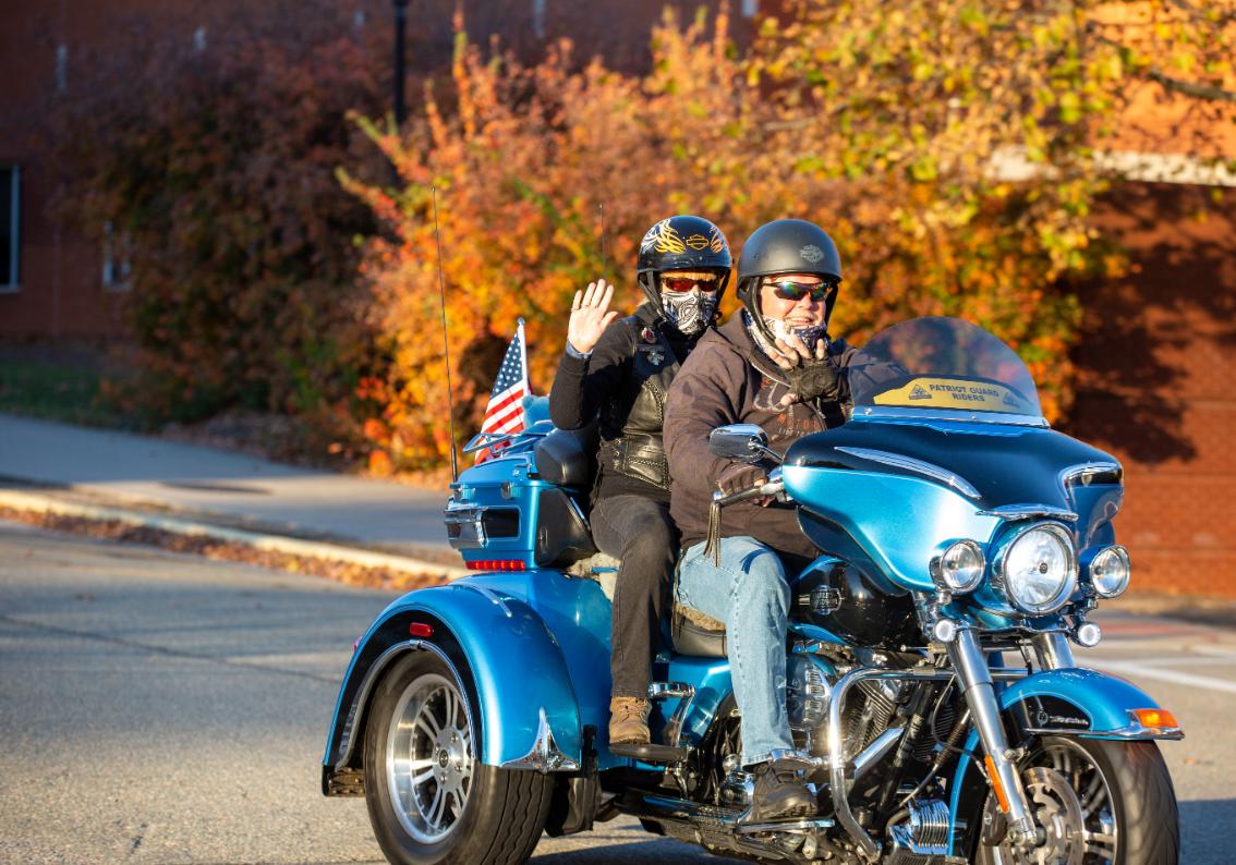 Mark and Allison Kendrick on Mark's motorcycle - Two riders cruise down a street on a blue three-wheeled motorcycle. Both wear helmets and sunglasses, and the passenger waves. An American flag is mounted behind the seat. Bright autumn leaves and a brick building fill the background.