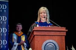 Dr. Kim Little - Woman in academic regalia smiles while speaking at a podium with the Longwood University seal. She stands on a stage in front of a microphone during a formal ceremony. A student in graduation attire applauds in the background. A Longwood University banner is visible beside the stage.