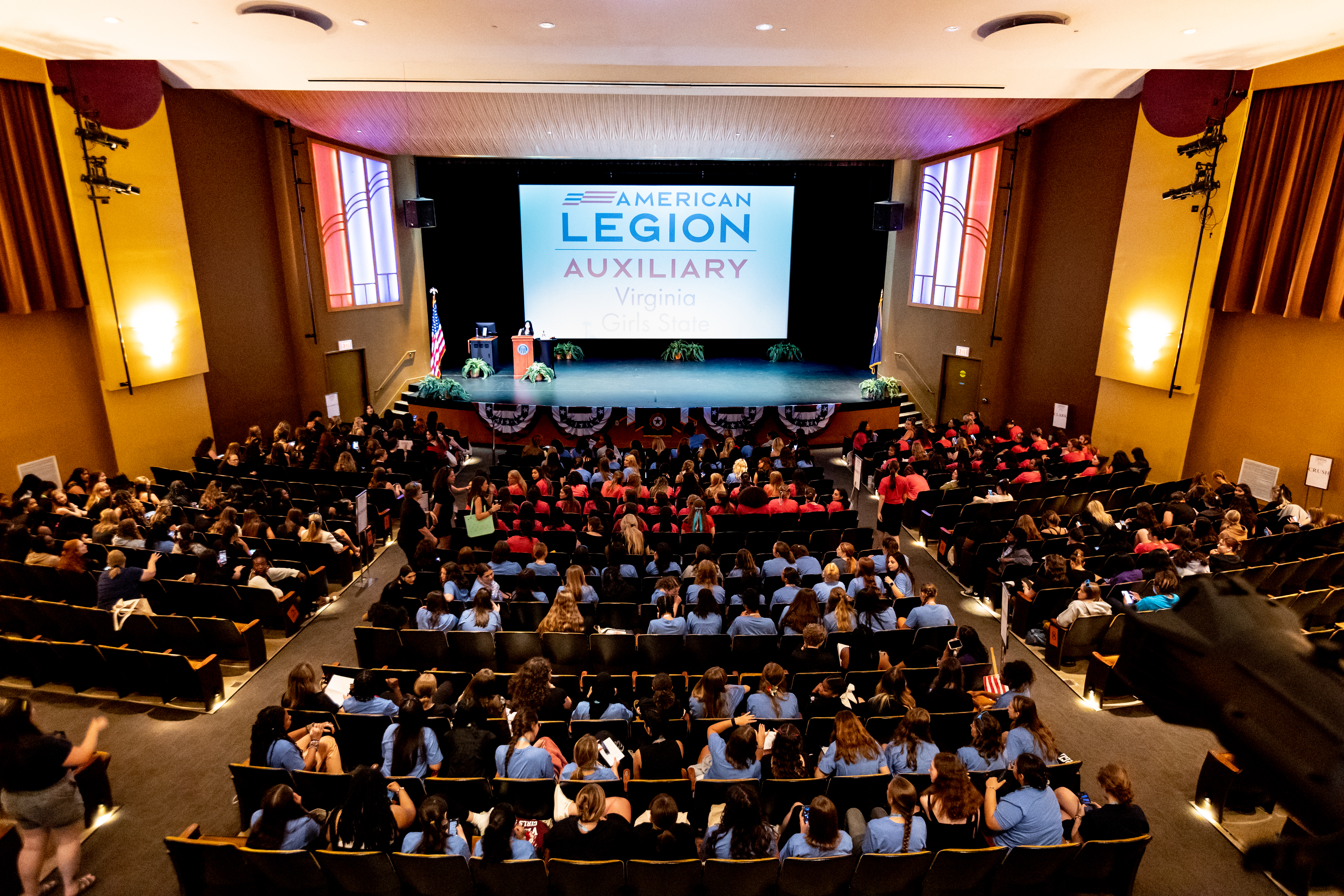 Girls State 2024 - Auditorium filled with attendees facing a stage at a formal event. Large screen reads “American Legion Auxiliary Virginia Girls State.” Audience members in blue and red shirts sit in tiered seating. Stage features a podium, flags, and patriotic bunting.  Is this conversation helpful so far?