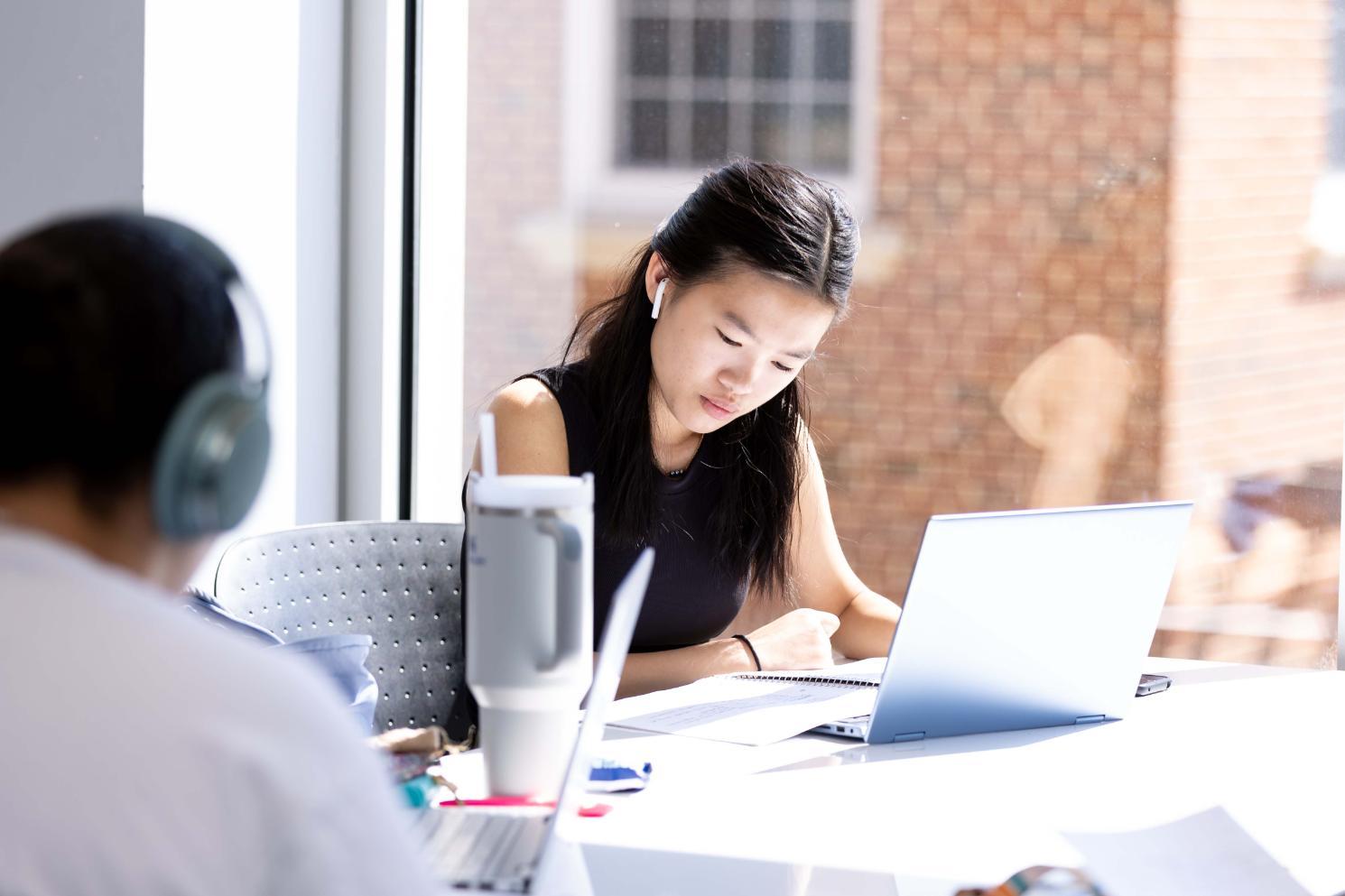 Student studying at laptop in the library - Student wearing wireless earbuds studies at a table near a large window. She looks down at a notebook beside an open laptop. Another student wearing headphones sits blurred in the foreground. Bright sunlight streams through the window, illuminating the study space.