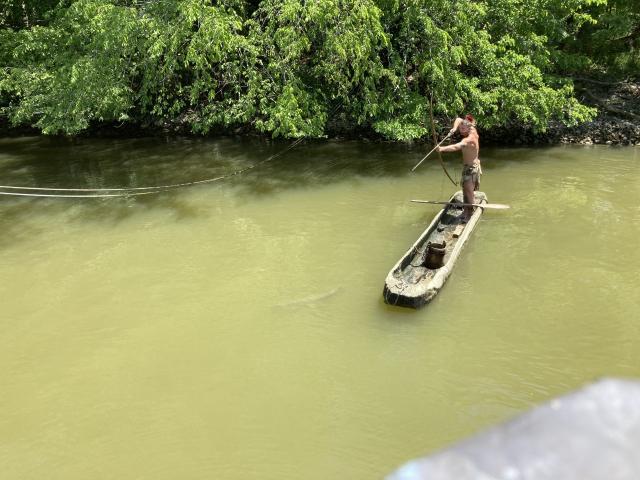 Russell Reed is standing in one of his hand-crafted canoes, drawing back on his bow and arrow to shoot a fish in the water.