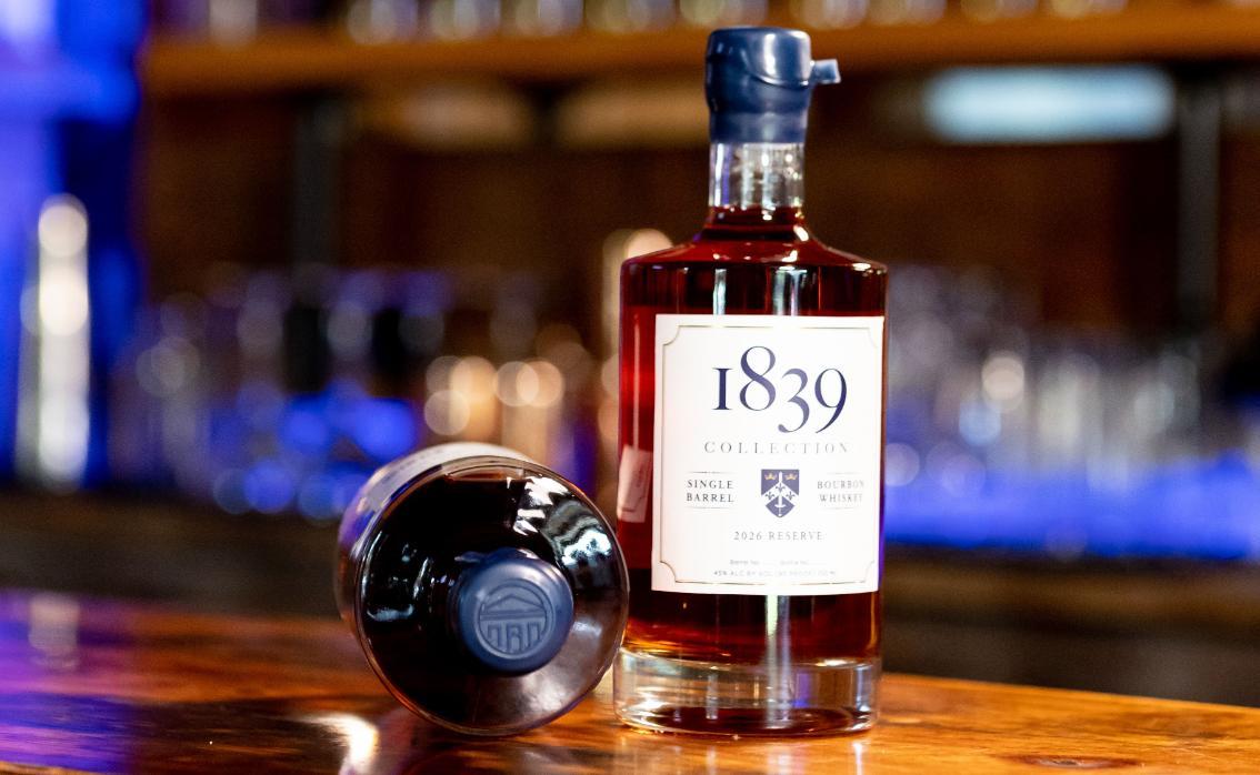 A bottle of Longwood’s 1839 Collection 2026 Reserve bourbon stands on a wooden bar beside a second bottle lying on its side, showing a blue cap embossed with the Longwood seal.