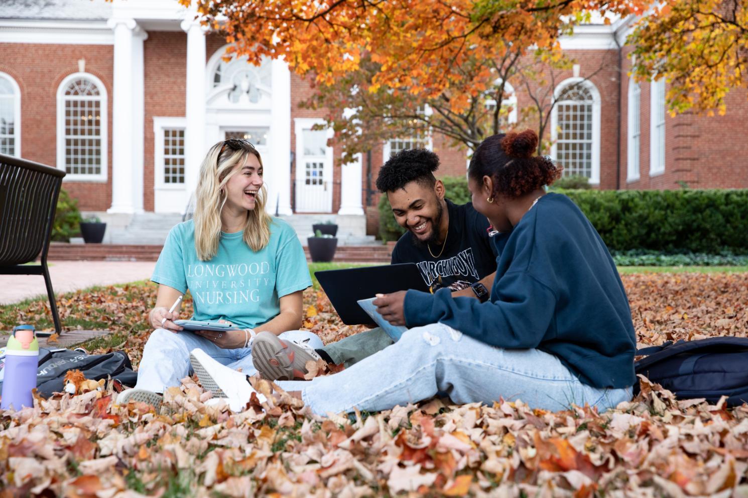Three students sit on the leaf-covered lawn in front of Eason Hall