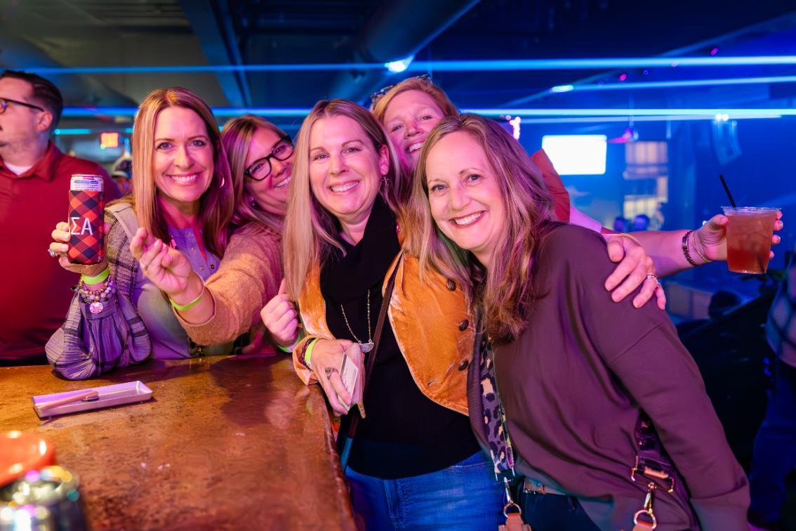 Julie Wiley Ramsey ’93 (second from right, pictured with classmates during an alumni event) describes herself as a 'proud townie' after moving back to Farmville from Virginia Beach in 2022. - Group of friends smiles and poses together at a bar under blue neon lighting. One person holds a drink can while another holds a cup with a straw. Arms are around each other as they lean against the bar count