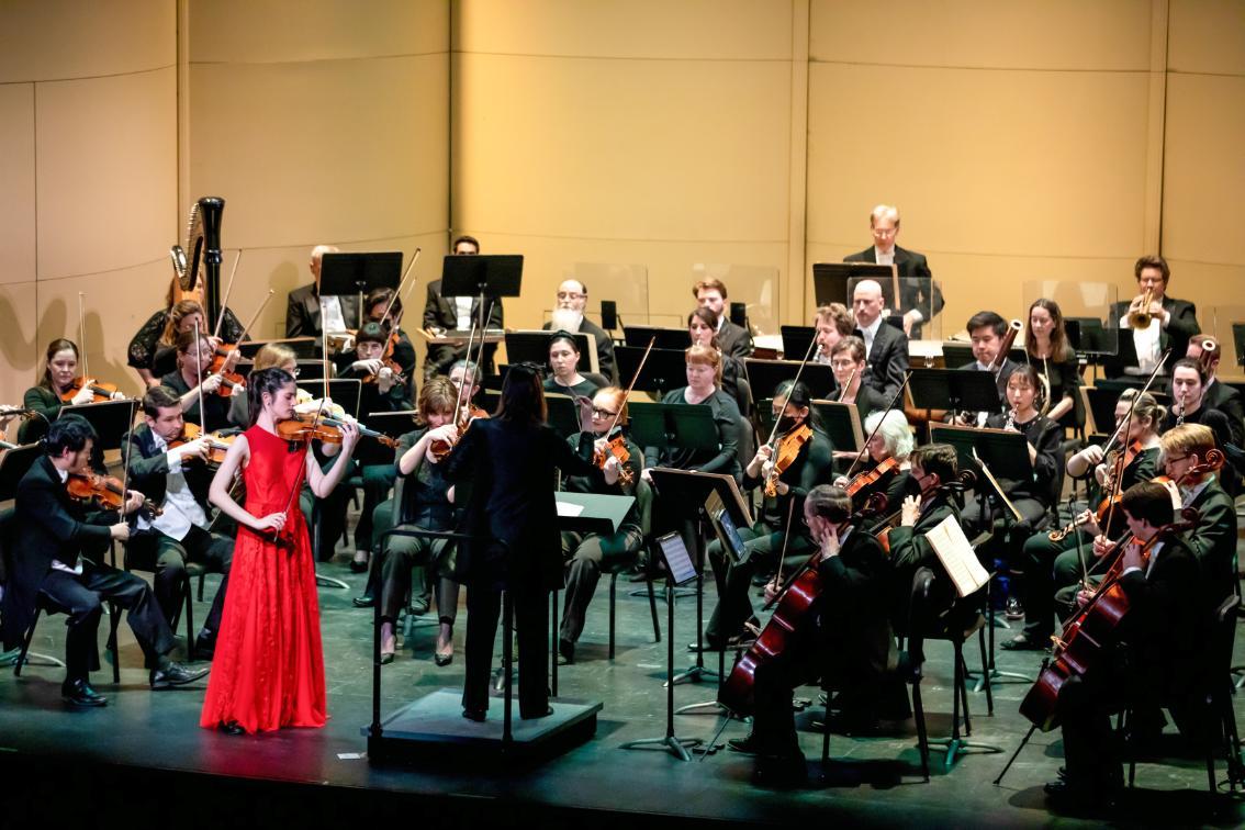 Richmond Symphony Orchestra performs on stage with a conductor leading and a violin soloist in a red gown at the front.