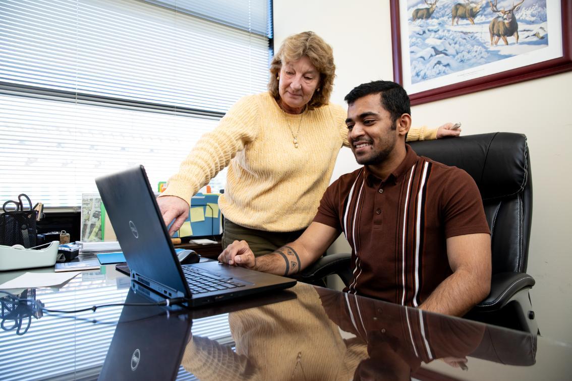 Two people collaborate at a desk while looking at a laptop in an office. A woman in a yellow sweater stands beside a seated man and points toward the screen. The man smiles while using the laptop at a glass-topped desk. Office items, window blinds, and a framed wildlife print hang on the wall behind them.