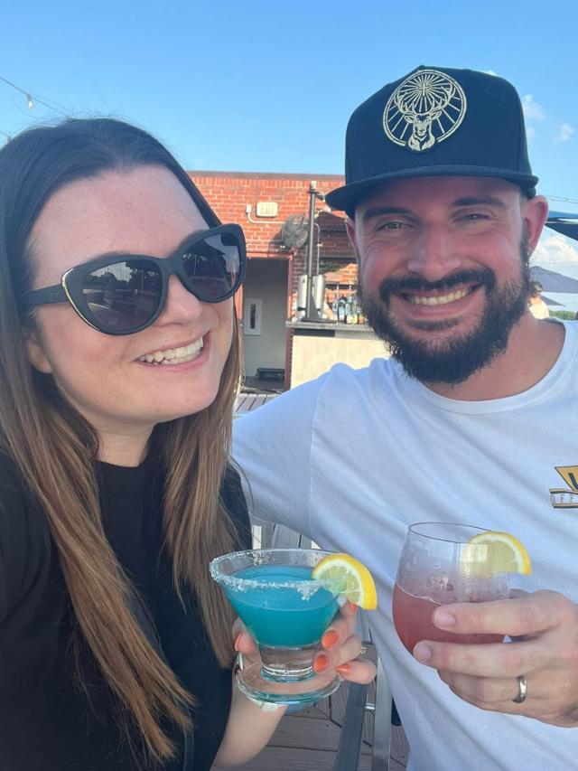 Keisha Stephens ’08 with her husband - Two people smile for a selfie outdoors at a rooftop patio bar. One wears sunglasses, and the other wears a black cap and has a beard. They hold colorful cocktails garnished with lemon slices. Brick wall and blue sky appear in the background.