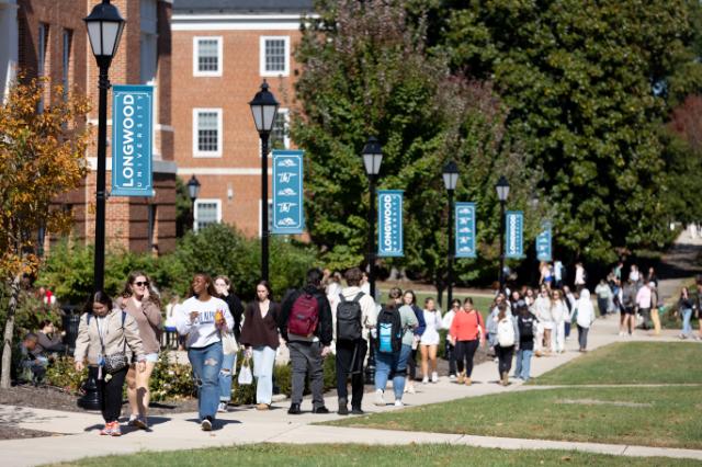 Students walk and talk along a busy campus sidewalk lined with Longwood banners on a sunny fall day.