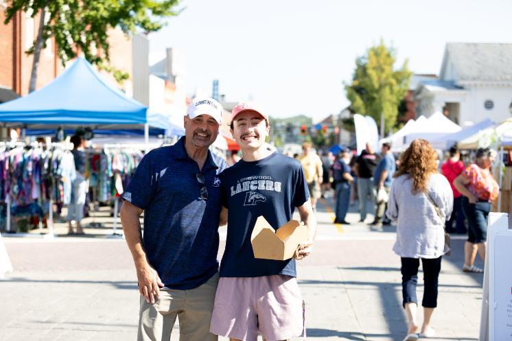 Two people smile together in Longwood gear during Family Weekend, with vendors and campus visitors in the background.